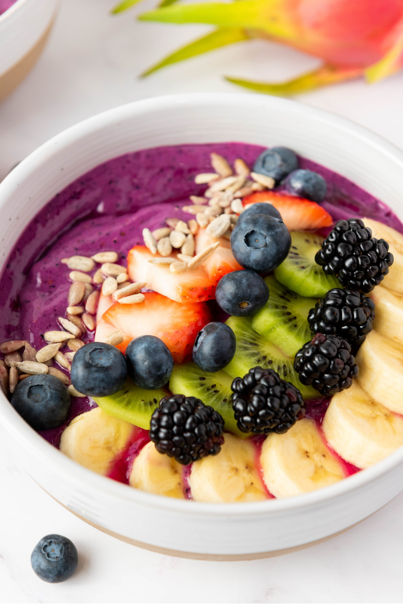 Thick purple smoothie bowl topped with sliced banana, kiwi, strawberries, blueberries, blackberries, and sunflower seeds in a white bowl.