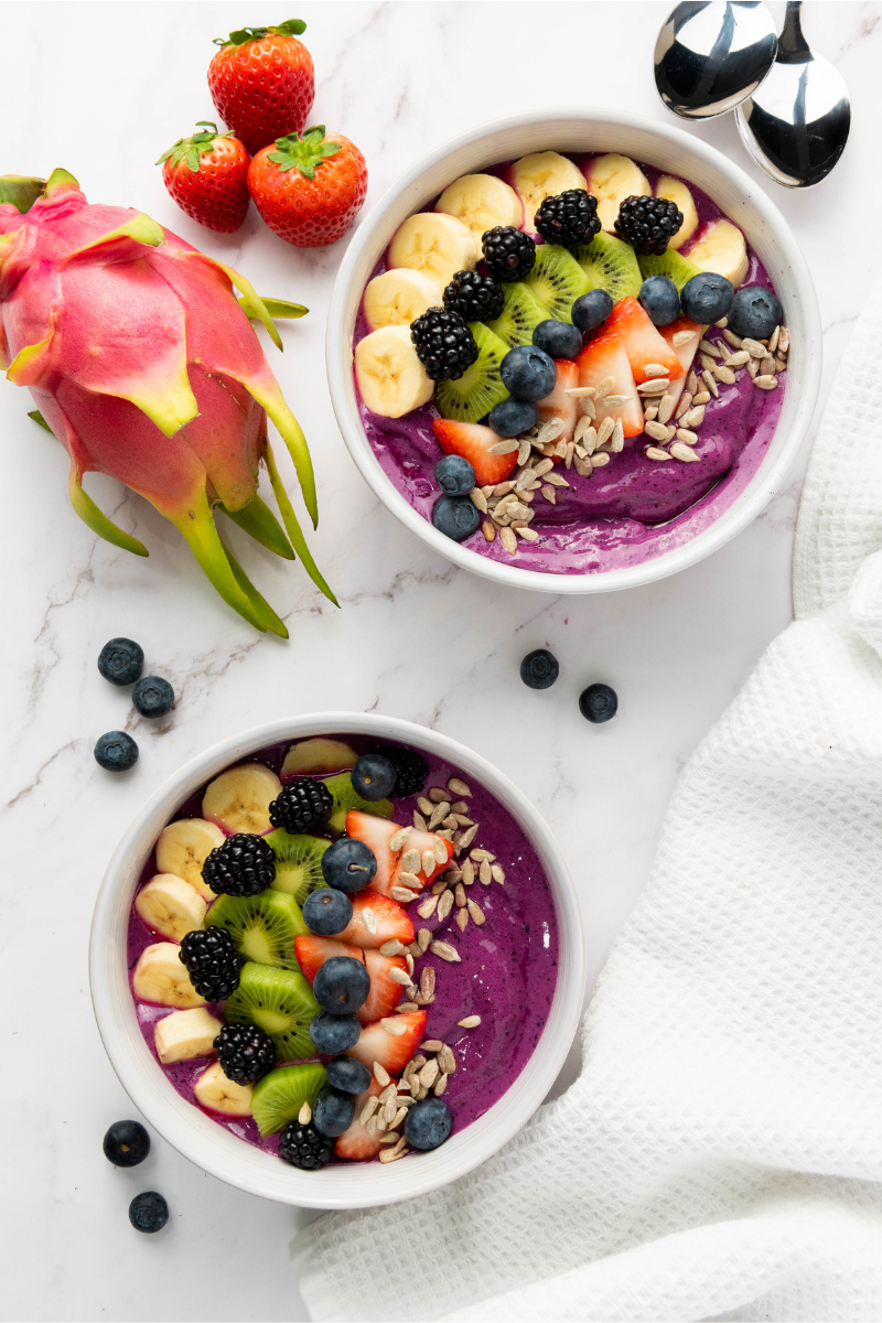 Two purple smoothie bowls topped with banana, kiwi, strawberries, blueberries, blackberries, and sunflower seeds on a marble surface.