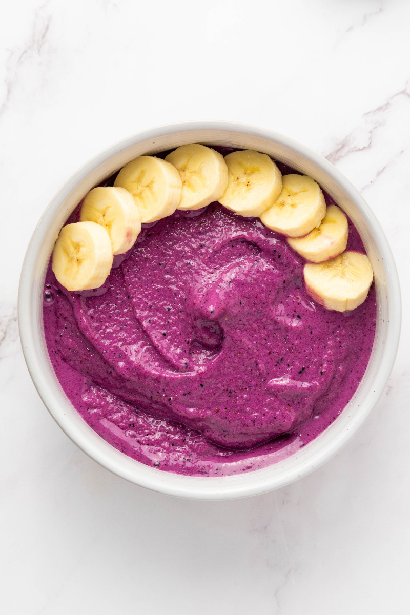 Purple smoothie bowl base topped with a row of sliced bananas as the first fruit topping layer.