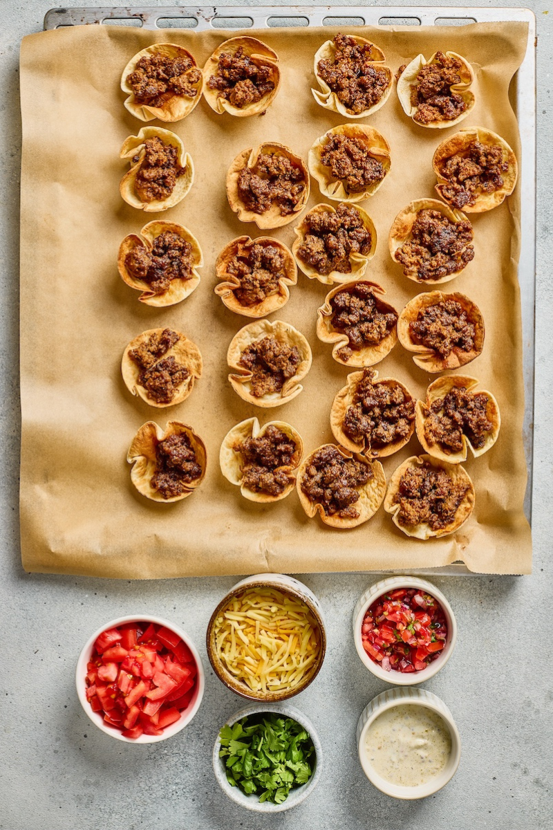 Crispy tortilla scoop cups filled with seasoned taco ranch beef on a parchment-lined baking sheet for an easy party appetizer.