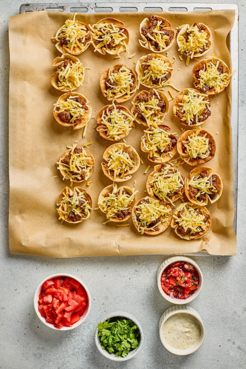 Taco ranch tortilla cups topped with shredded cheese before baking, with salsa, cilantro, tomatoes, and ranch sauce on the side.