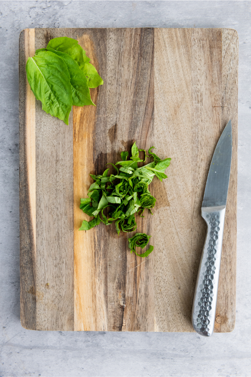 Fresh basil sliced on a wooden cutting board with a kitchen knife for topping mini Caprese naan pizzas.