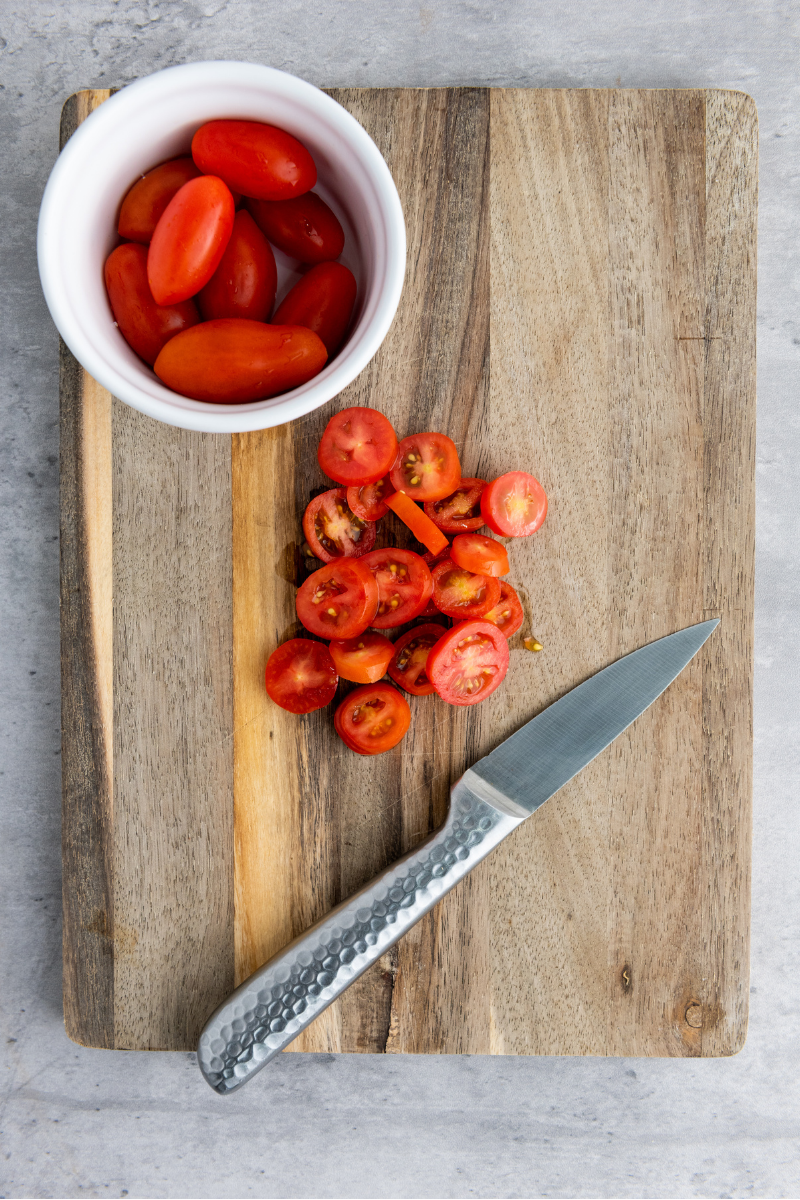 Cherry tomatoes sliced on a wooden cutting board with a kitchen knife for preparing mini Caprese naan pizzas.