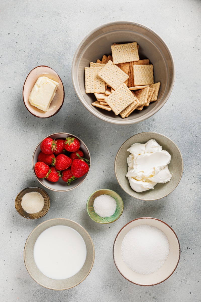 Ingredients for no-bake strawberry cheesecake including strawberries, cream cheese, sugar, gelatin, cream, butter, and graham crackers arranged on a countertop.
