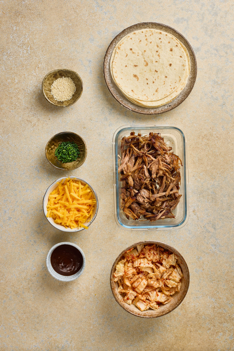 Quick kimchi mixed with chili seasoning beside pulled pork, tortillas, cheese, sesame seeds, green onions, and barbecue sauce.