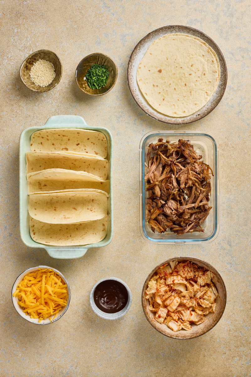 Small tortillas arranged upright in a baking dish with pulled pork, quick kimchi, cheddar cheese, sesame seeds, green onions, and barbecue sauce nearby.