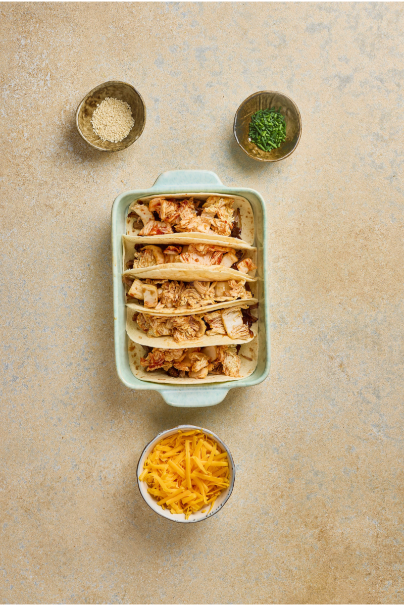 Quick kimchi spooned over pulled pork tacos in a baking dish with cheddar cheese ready to add.

