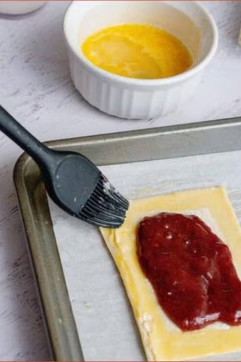 Egg wash being brushed onto raspberry jam turnovers before baking until golden brown