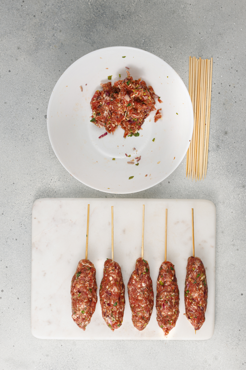 Kofte mixture being shaped around wooden skewers into traditional elongated kebabs.