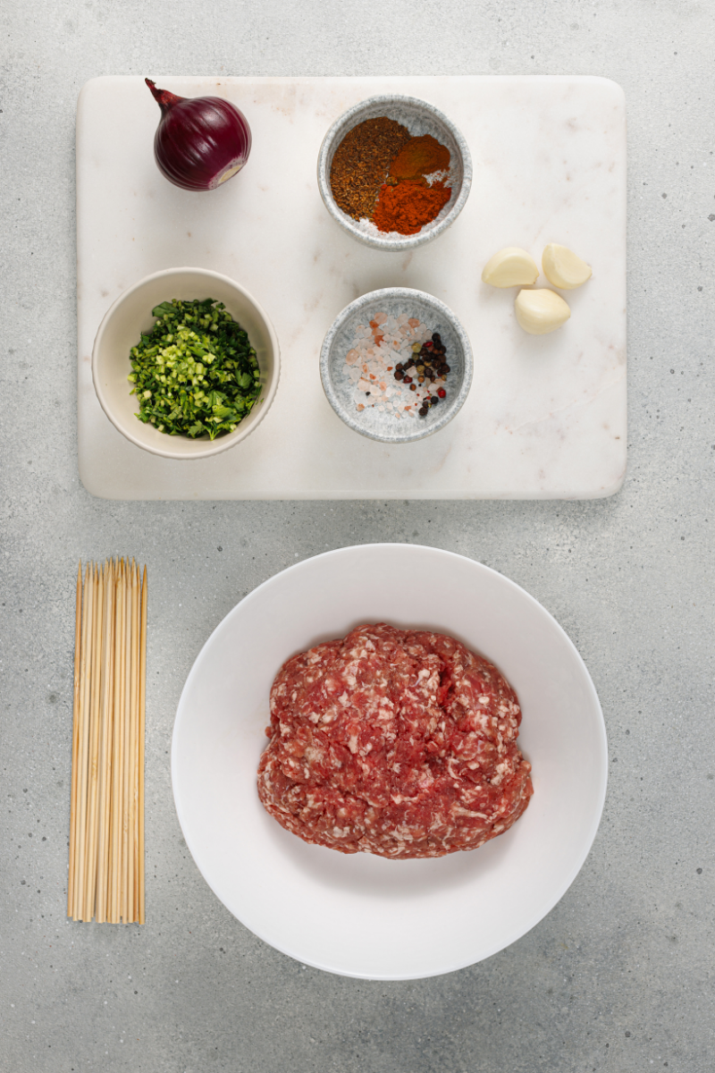 Ground meat, onion, garlic, parsley, spices, olive oil, and skewers arranged on a countertop before preparing homemade kofte kebabs.