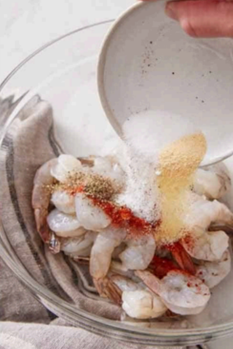 Adding garlic powder, paprika, salt, and black pepper to raw shrimp in a glass bowl before air frying.