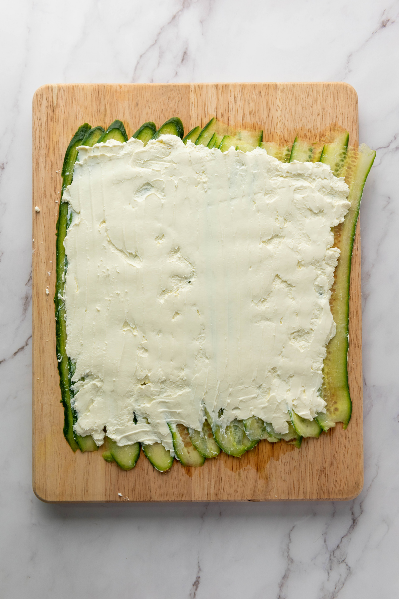 Spreading softened cream cheese evenly over cucumber ribbons for Everything Bagel Cucumber Rolls.