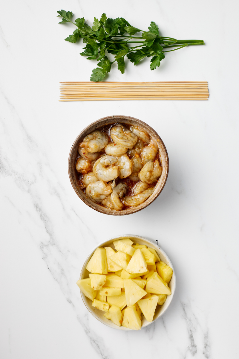 Marinated shrimp and fresh pineapple chunks in bowls with wooden skewers ready to assemble pineapple shrimp skewers
