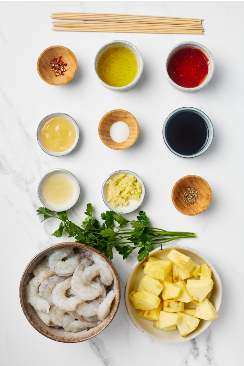Flat lay of all ingredients for sweet and spicy pineapple shrimp skewers — raw shrimp, pineapple chunks, olive oil, soy sauce, honey, garlic, ginger, chili flakes, and wooden skewers