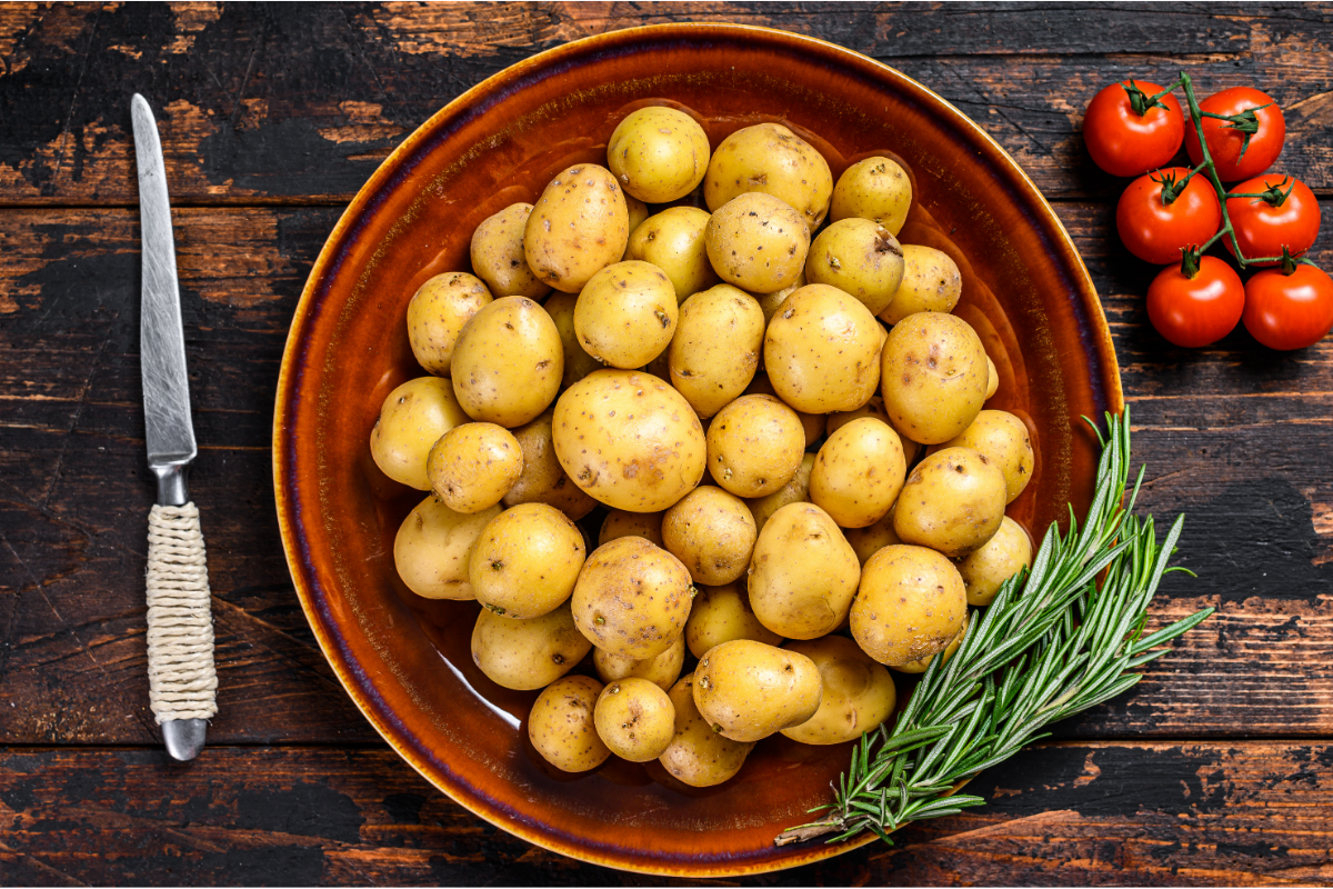 Baby yellow potatoes in a brown bowl with tomatoes, rosemary and a kitchen knife.