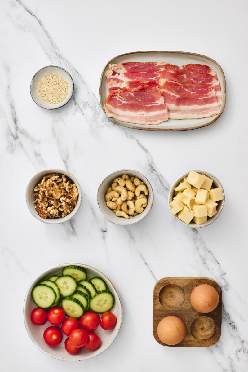  Ingredients for savory protein lunchboxes including eggs, bacon, cheese, walnuts, cashews, cucumber, cherry tomatoes, and sesame seeds.