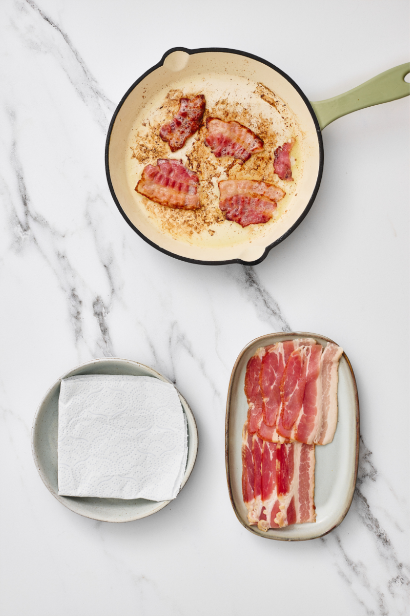 acon cooking in a skillet with uncooked bacon and paper towels nearby.

