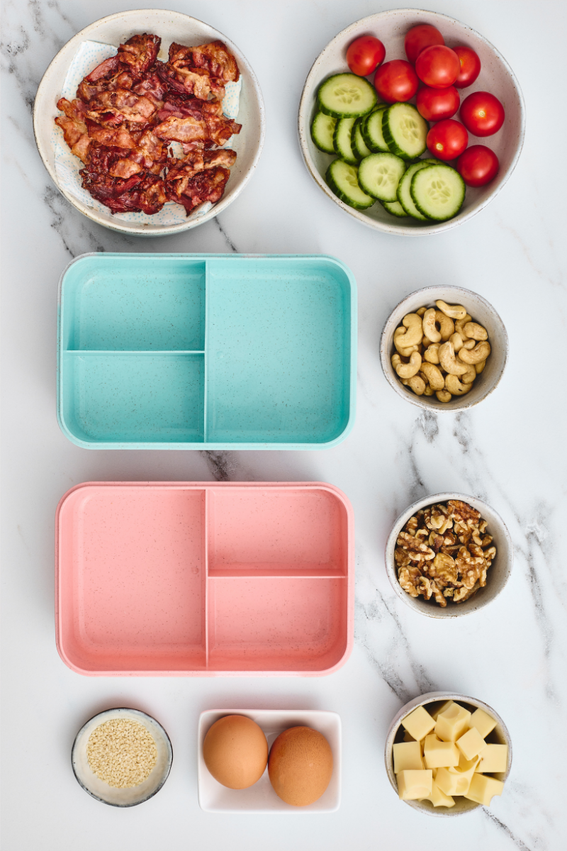 Two empty compartment lunchboxes surrounded by cooked bacon, eggs, cheese, walnuts, cashews, cucumber, tomatoes, and sesame seeds.

