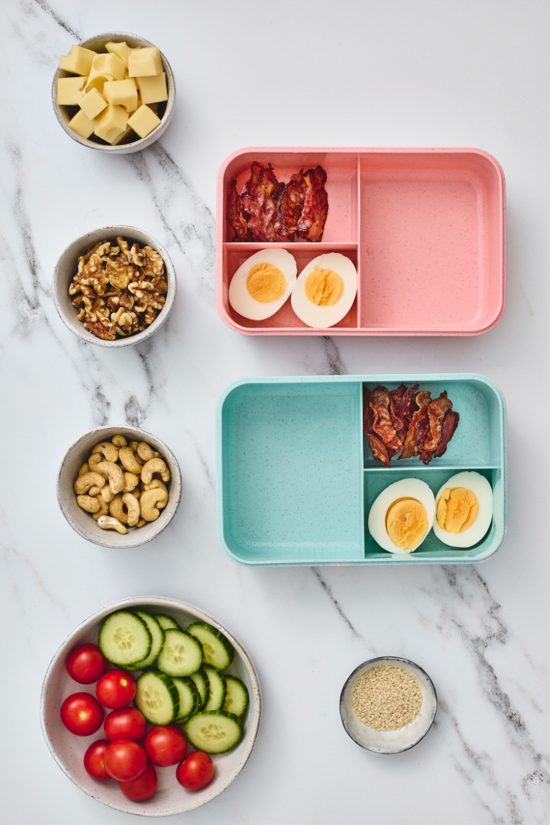 Crispy bacon and hard-boiled egg halves arranged in two compartment lunchboxes.