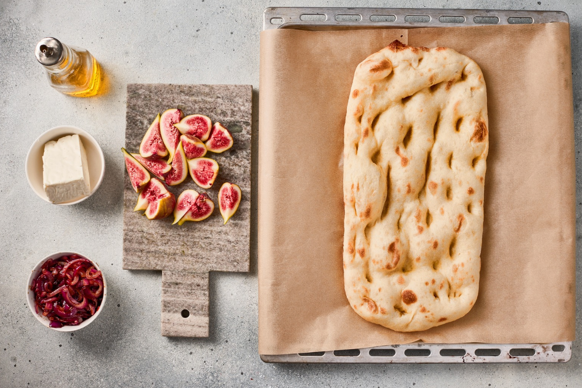Fresh figs cut into wedges beside a prepared rectangular flatbread on a parchment-lined baking sheet.