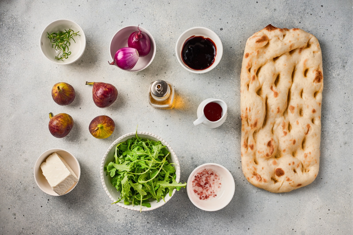 Overhead arrangement of figs, red onions, flatbread, white cheese, arugula, rosemary, olive oil, balsamic sauce, and salt for fig flatbread.