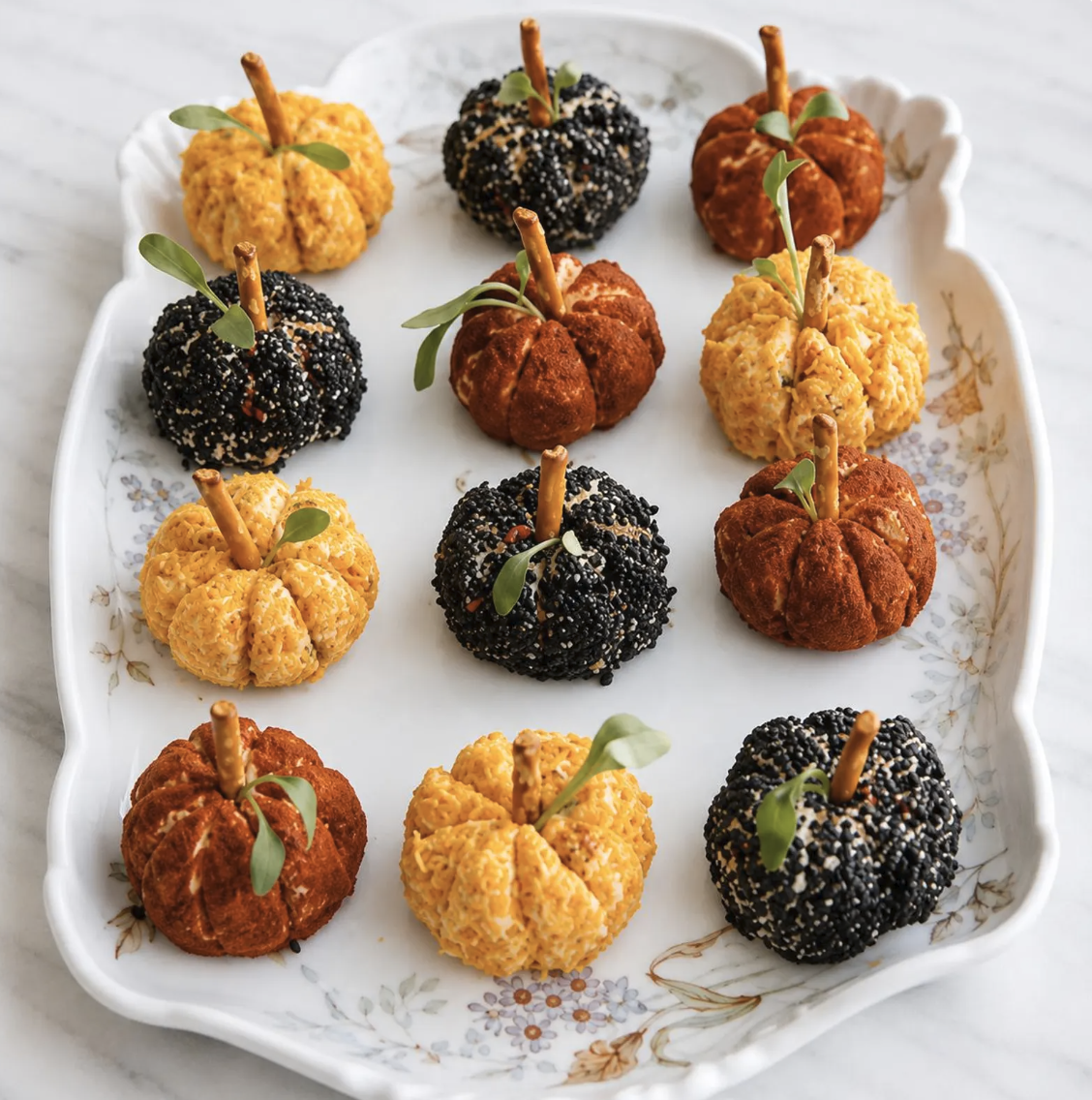 Finished orange, red, and black mini pumpkin cheese balls with pretzel stems and fresh green leaves arranged on a platter for a festive Fall, Autumn, and Thanksgiving appetizer.
